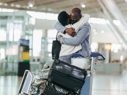 A couple embracing in an airport arrivals hall beside a loaded luggage trolley.