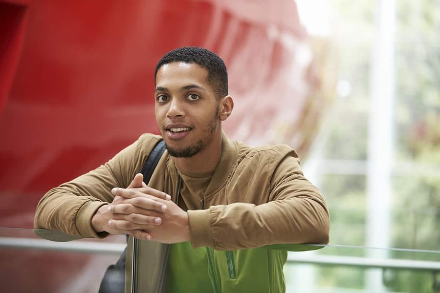 A recent graduate leaning on a glass balcony rail inside a modern university building.