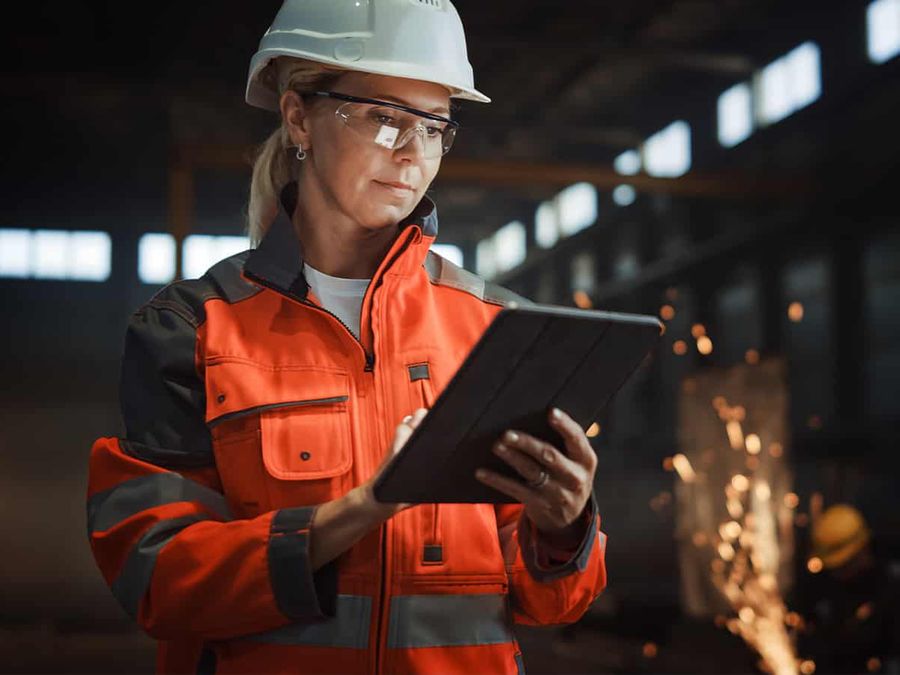 An engineer in a hard hat and orange hi-vis jacket checking a tablet on a factory floor.