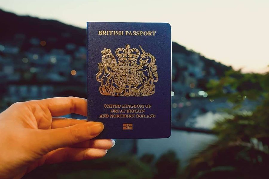 A hand holding up a blue British passport in front of a harbour town at sunset.