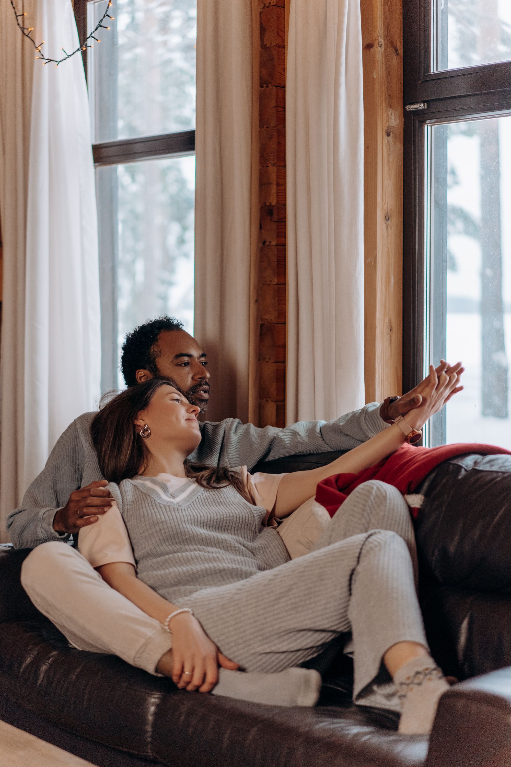 A couple curled up together on a sofa in a wood-framed living room, hands clasped, looking out at a snowy view - the settled life you are building here in the UK is what the partner and spouse visa is designed to protect.