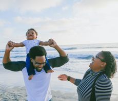 A father on the beach lifts his baby overhead as the mother laughs beside them - the settled family life the UK partner and family routes are built to protect.